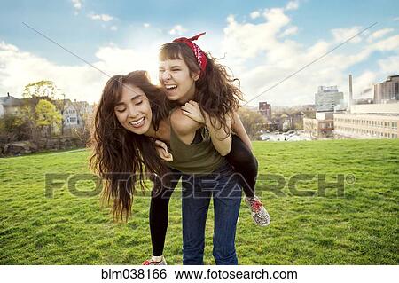 Woman carrying friend piggyback in urban park View Large Photo Image Stock Photograph - Woman carrying friend piggyback in urban park. Fotosearch