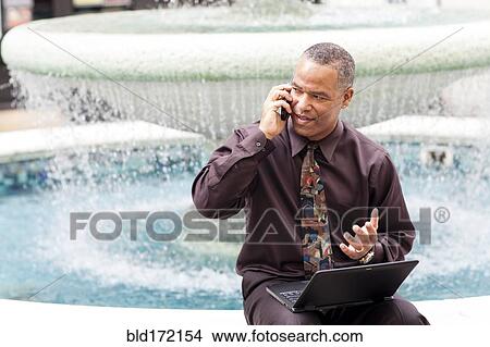 Picture - African American businessman talking on cell phone at fountain. Fotosearch