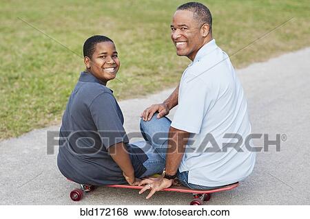 Stock Photo - African American father and son sitting on skateboard on path. Fotosearch