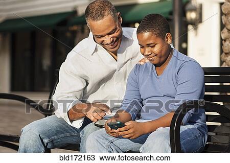 Stock Image - African American father and son using cell phone on bench. Fotosearch