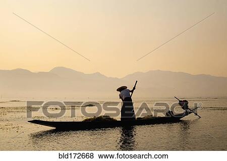 Stock Photo - Asian fisherman rowing canoe on rural lake. Fotosearch