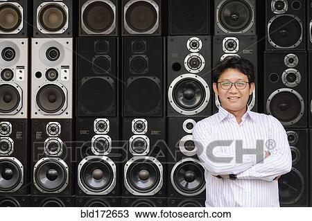 Stock Image - Asian man standing by stacked speakers. Fotosearch