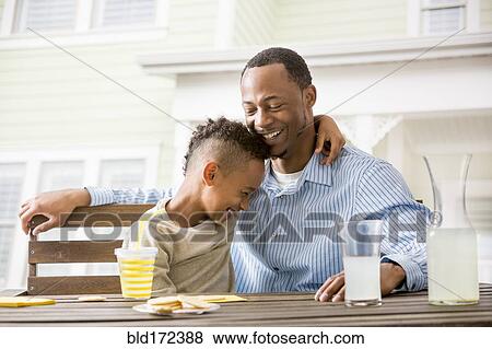 Stock Photo - Black father and son hugging at table in backyard. Fotosearch