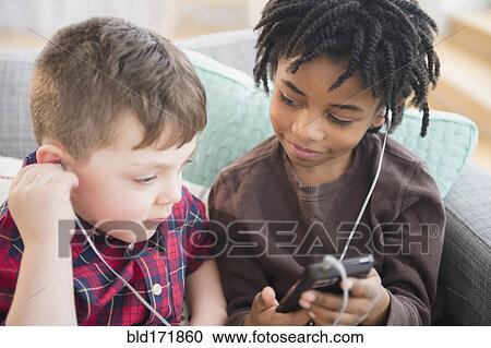 Stock Image - Boys sharing headphones and cell phone on sofa. Fotosearch