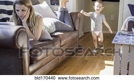 Stock Image - Caucasian baby boy walking past mother on sofa. Fotosearch