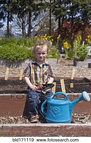 Stock Photo - Caucasian boy holding spade with watering can in garden. Fotosearch