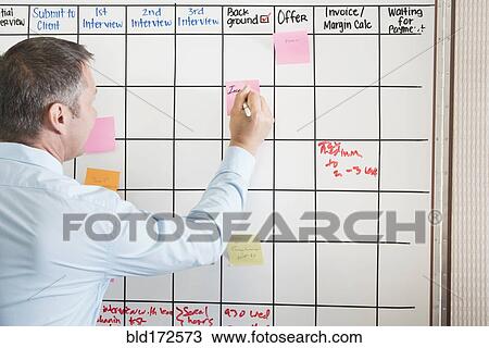 Stock Image - Caucasian businessman writing sticky note on office calendar. Fotosearch