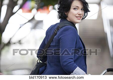 Stock Image - Caucasian businesswoman pushing bicycle outdoors. Fotosearch