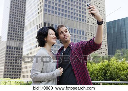 Stock Image - Caucasian couple taking selfie with cell phone in city. Fotosearch