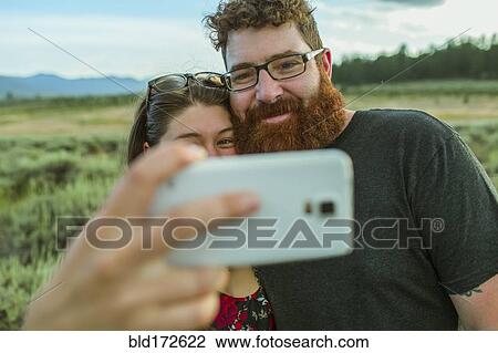 Stock Image - Caucasian couple taking selfie with cell phone in remote field. Fotosearch