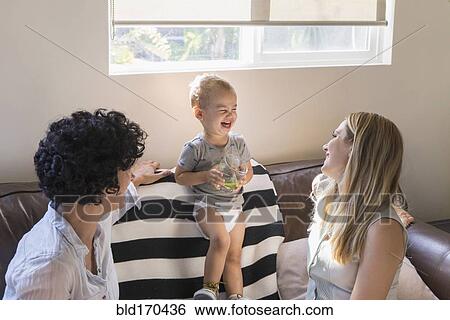 Stock Photograph - Caucasian lesbian mothers laughing with baby son on sofa. Fotosearch