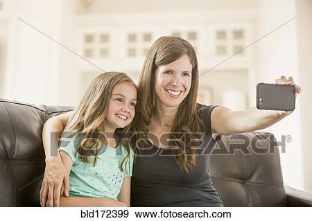 Stock Photo - Caucasian mother and daughter taking selfie with cell phone on sofa. Fotosearch
