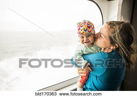 Stock Image - Caucasian mother kissing baby daughter at window. Fotosearch