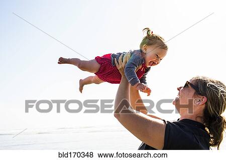 Stock Photo - Caucasian mother lifting baby daughter on beach. Fotosearch