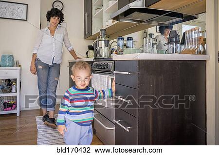 Stock Image - Caucasian mother watching baby son in kitchen. Fotosearch