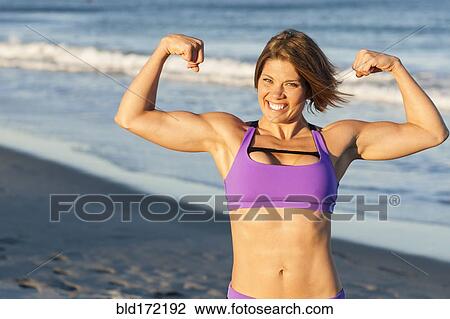 Stock Image - Caucasian woman flexing muscles on beach. Fotosearch