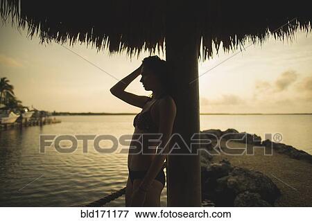Stock Photo - Caucasian woman standing under thatch umbrella on beach. Fotosearch