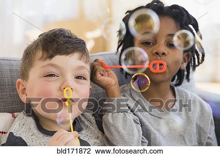 Stock Image - Close up of boys blowing bubbles on sofa. Fotosearch