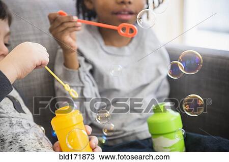 Stock Image - Close up of boys blowing bubbles on sofa. Fotosearch