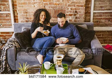 Picture - Couple eating breakfast on sofa in living room. Fotosearch