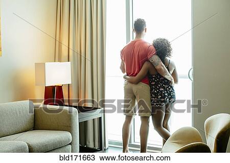 Stock Image - Couple hugging and looking out hotel window. Fotosearch