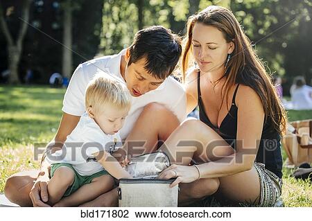 Stock Image - Family opening picnic lunch box in park. Fotosearch