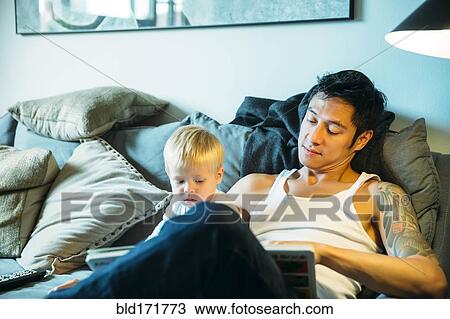 Stock Image - Father and son reading on sofa in living room. Fotosearch