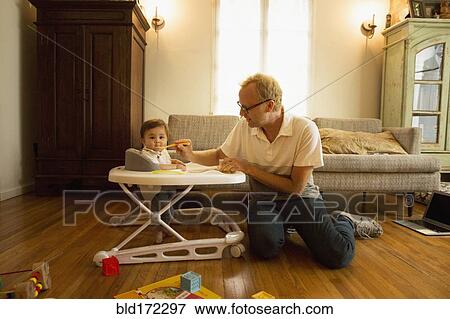 Father feeding son in high chair in living room View Large Photo Image Stock Photo - Father feeding son in high chair in living room. Fotosearch