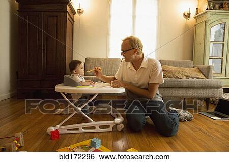 Stock Photograph - Father feeding son in high chair in living room. Fotosearch