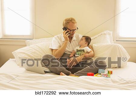Stock Image - Father holding son and talking on cell phone on bed. Fotosearch