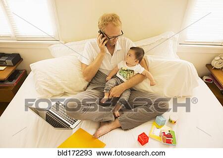 Father holding son and talking on cell phone on bed View Large Photo Image Stock Image - Father holding son and talking on cell phone on bed. Fotosearch