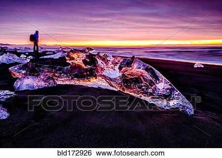 Stock Photograph - Hiker admiring glaciers on beach reflecting sunset sky. Fotosearch