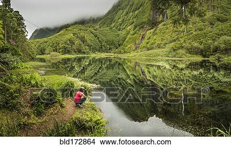 Picture - Hiker crouching at still lake in remote mountains. Fotosearch