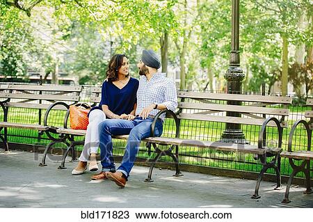 Stock Image - Indian couple sitting on bench in urban park. Fotosearch