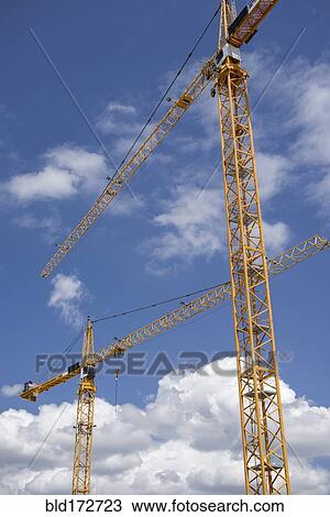 Stock Image - Low angle view of construction cranes under blue sky. Fotosearch