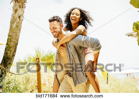 Picture - Man carrying girlfriend piggyback on beach. Fotosearch