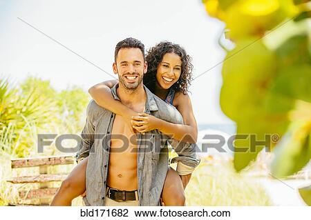 Stock Image - Man carrying girlfriend piggyback on beach. Fotosearch