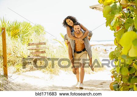 Stock Image - Man carrying girlfriend piggyback on beach. Fotosearch