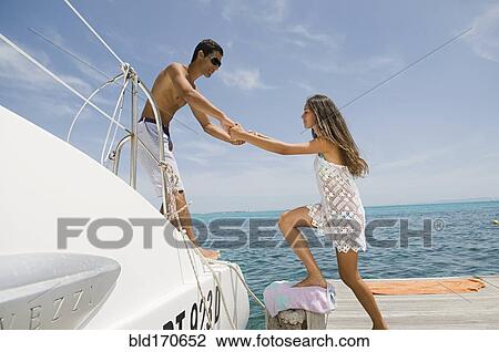 Stock Image - Man helping girlfriend into sailboat on harbor. Fotosearch