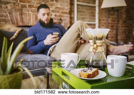 Stock Image - Man using cell phone with breakfast in living room. Fotosearch