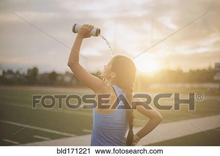 Stock Image - Mixed race athlete pouring water bottle on herself on sports field. Fotosearch