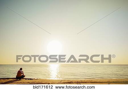 Stock Image - Mixed race man sitting on beach. Fotosearch