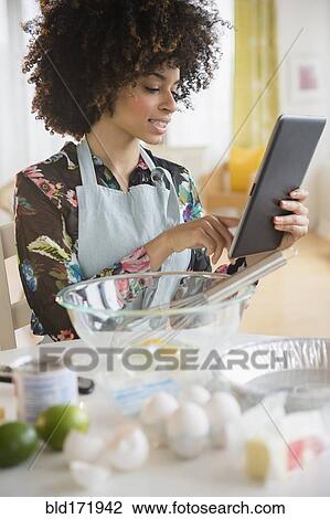 Stock Image - Mixed race woman baking with recipe on digital tablet. Fotosearch