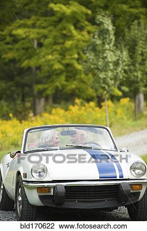 Stock Image - Older couple driving convertible on dirt road. Fotosearch