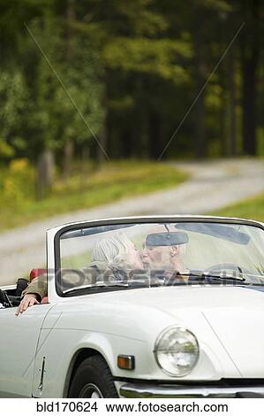 Picture - Older couple kissing in convertible on dirt road. Fotosearch