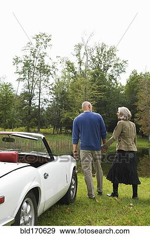 Stock Photo - Rear view of couple holding hands by convertible in park. Fotosearch