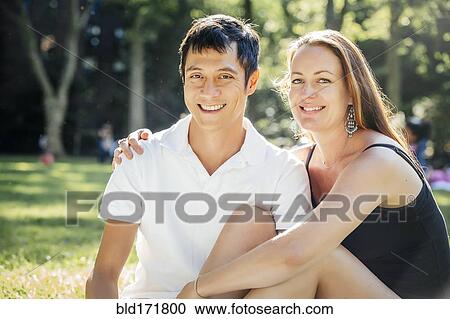 Stock Image - Smiling couple sitting on grass in park. Fotosearch