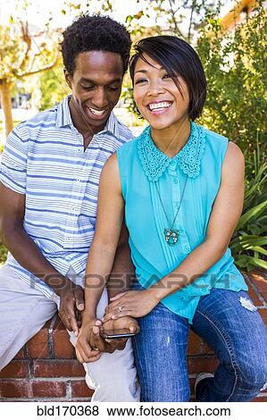 Stock Photo - Smiling couple using cell phone outdoors. Fotosearch