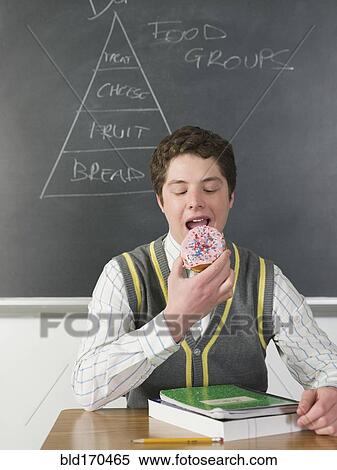 Stock Photography - Student eating donut at chalkboard in classroom. Fotosearch