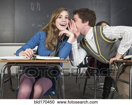 Stock Image - Students whispering at desks in classroom. Fotosearch
